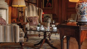 A classic library setting with a large armchair, a coffee table, and a bookshelf filled with books. - Lucknam Park, Emblems Collection (Chippenham)