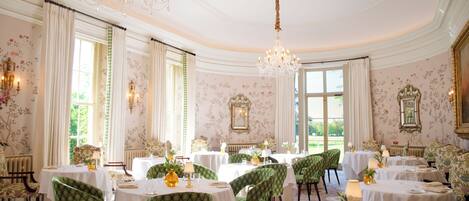 Un comedor formal con mesas dispuestas para el servicio, sillas tapizadas de verde y candelabros ornamentados.