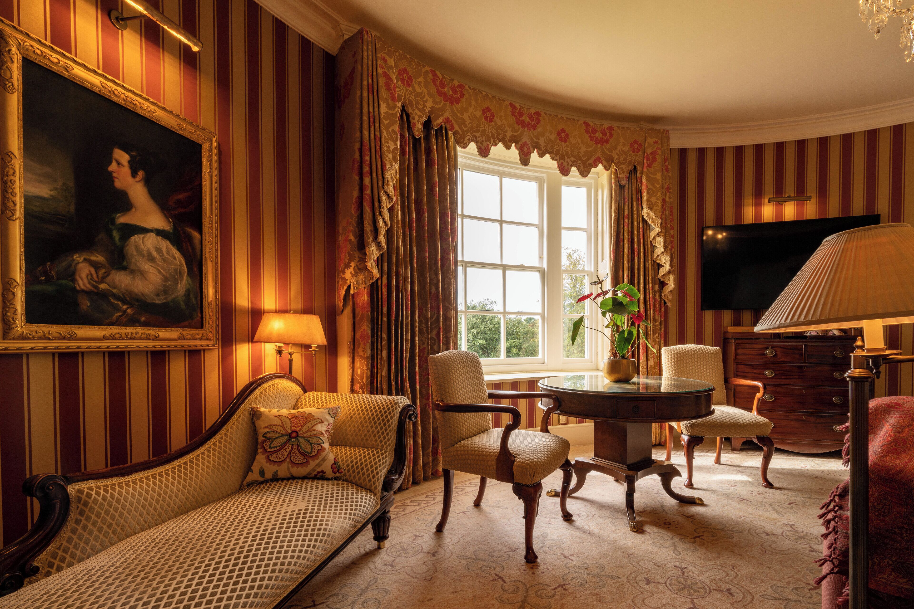 A room with striped wallpaper, a large painting, a sofa, a dining table with chairs, a television, and a window with curtains.