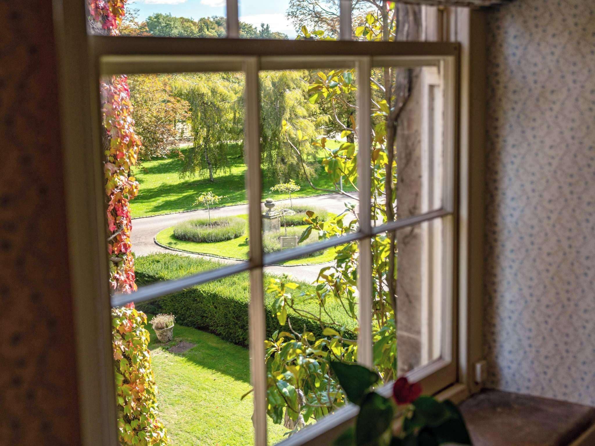 A window with a view of a well-maintained garden with a curved pathway, neatly trimmed hedges, and a small statue.