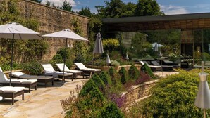A patio area with lounge chairs and umbrellas, surrounded by greenery and a stone wall. - Lucknam Park, Emblems Collection (Chippenham)