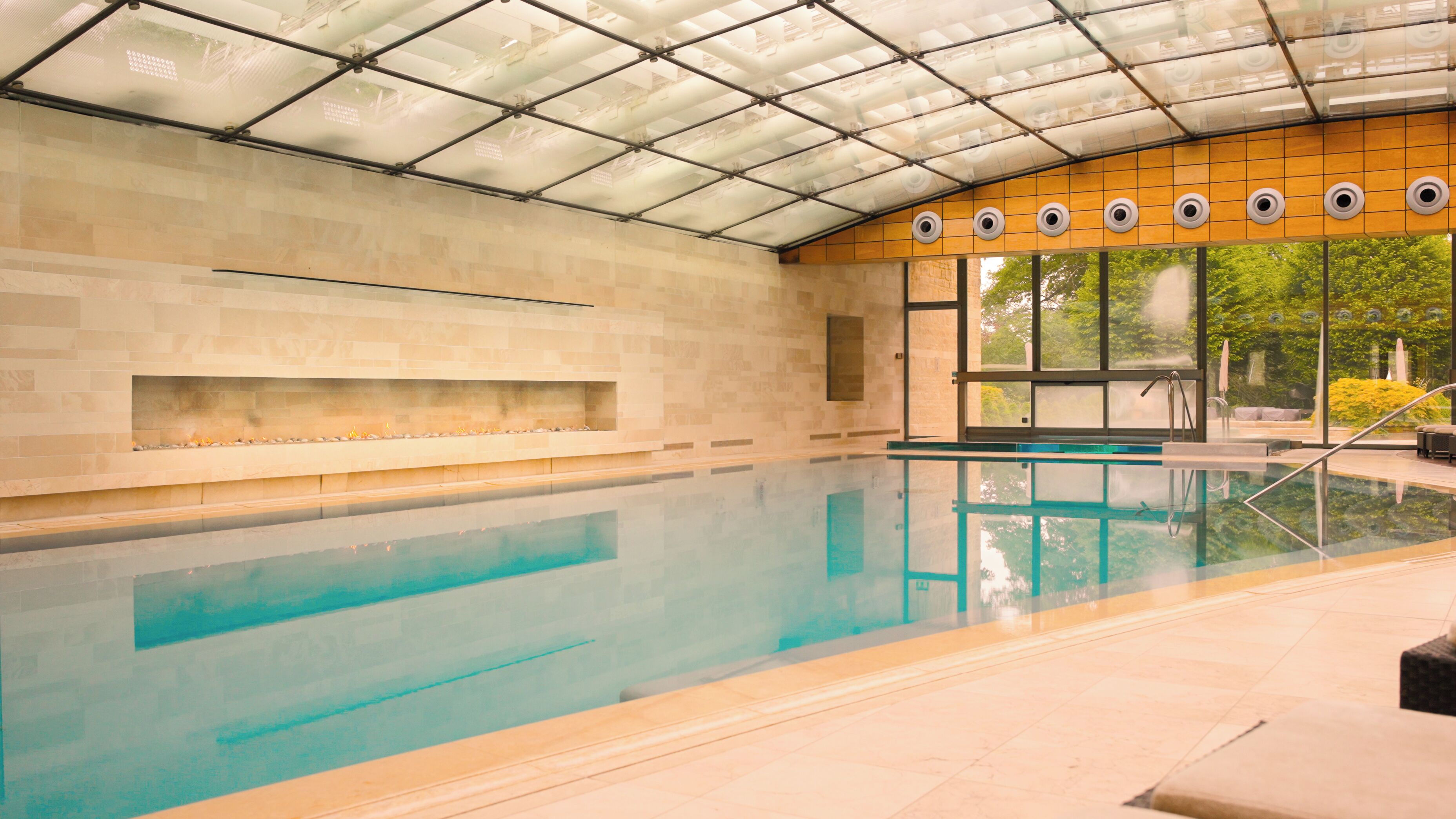 An indoor swimming pool with a glass ceiling, stone walls, and a view of greenery outside.