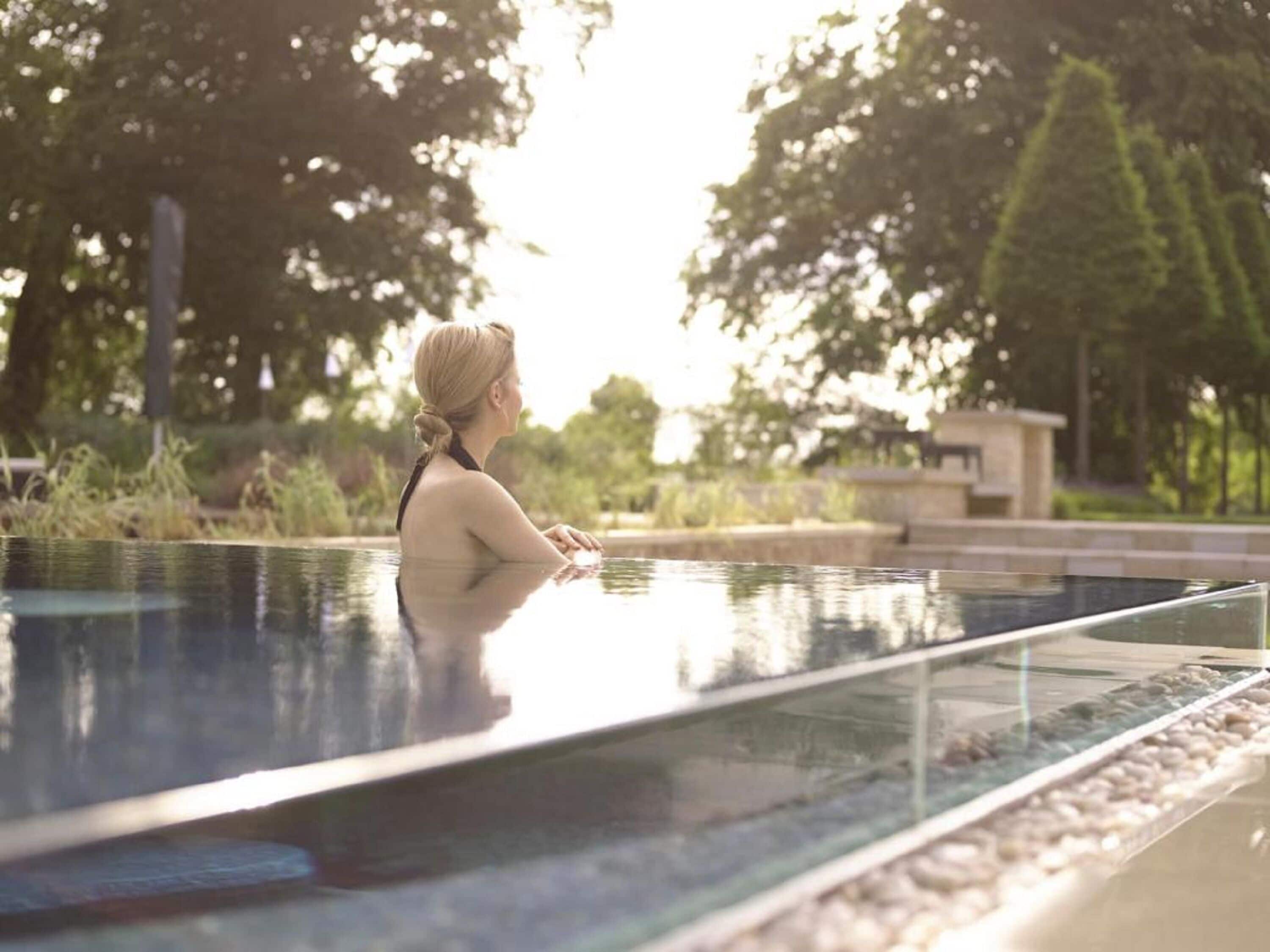 Une personne se détend dans une piscine aux eaux cristallines, avec en arrière-plan des arbres et un petit bâtiment.