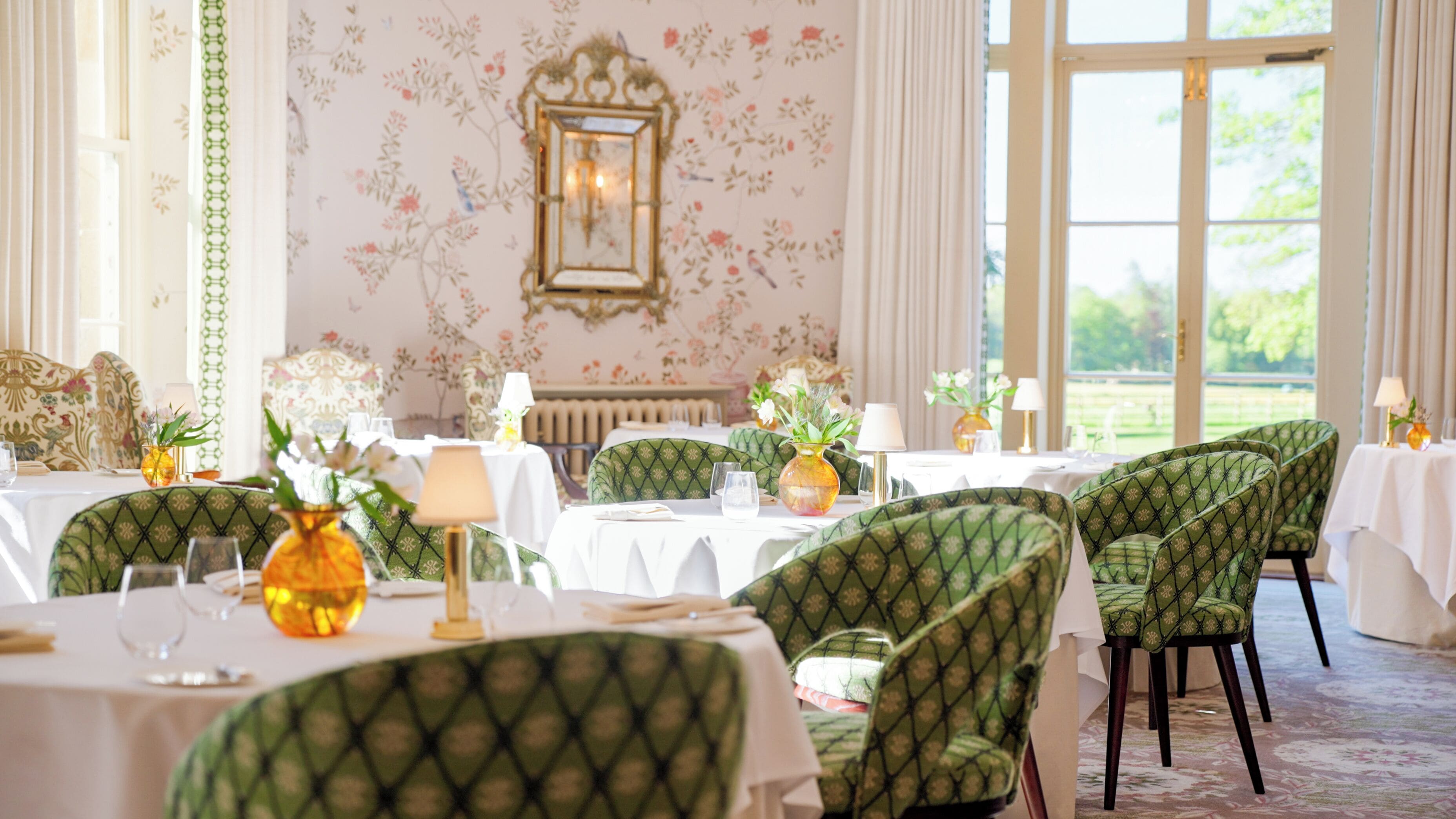 A dining area with green upholstered chairs, white tablecloths, and floral wallpaper.