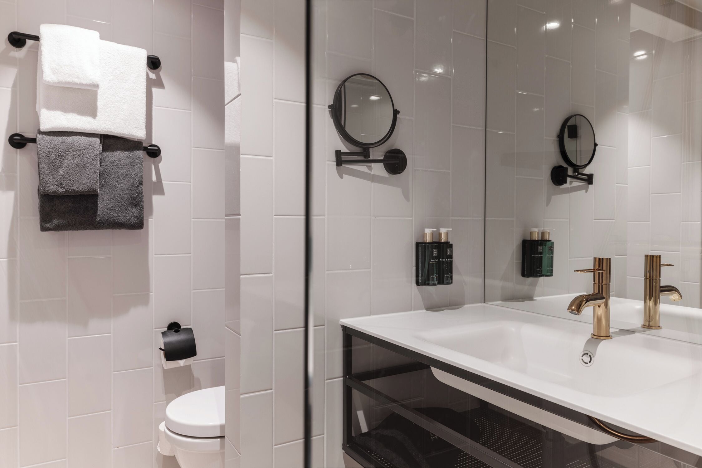 Un baño moderno con un lavamanos blanco, grifos dorados y dos espejos montados en la pared. Hay toallas en un perchero, un dispensador de papel higiénico y una zona de ducha con mampara de cristal.