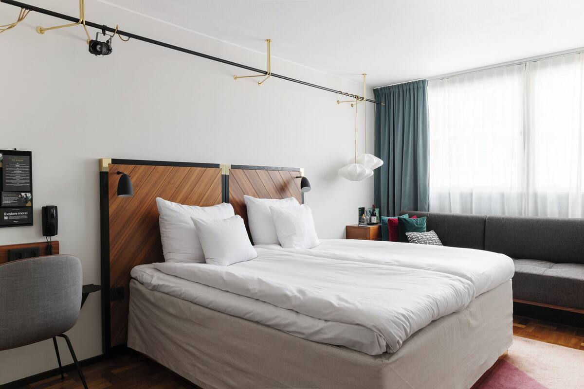 Quarto de hotel moderno com uma cama grande, um sofá, uma escrivaninha e um projetor no teto.