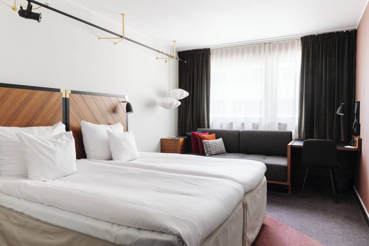 Quarto de hotel com uma cama grande, uma escrivaninha, uma cadeira, um sofá e uma janela com cortinas.