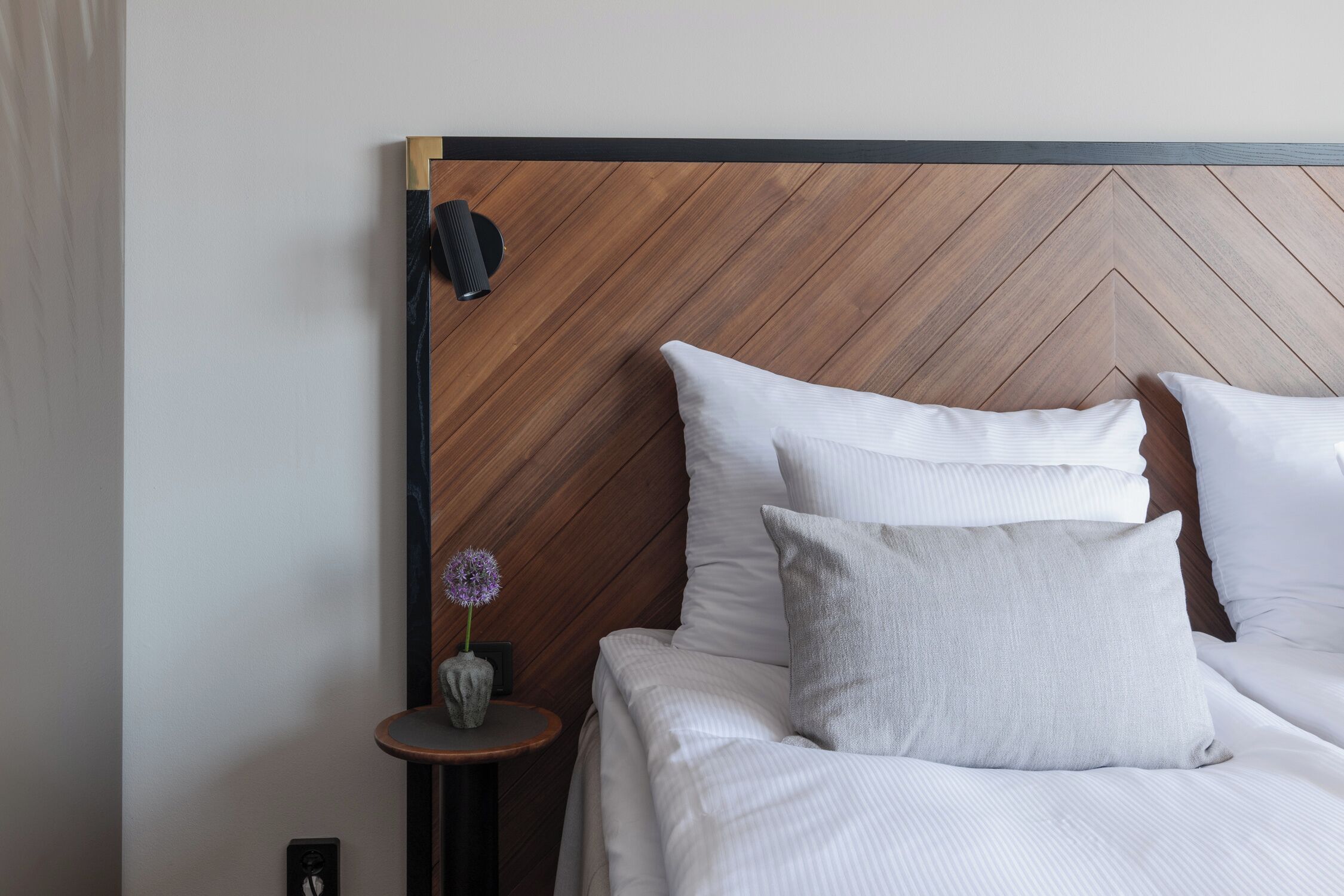 A neatly made bed with white linens and two pillows, a small round table with a vase of flowers, and a wooden headboard with a chevron pattern.