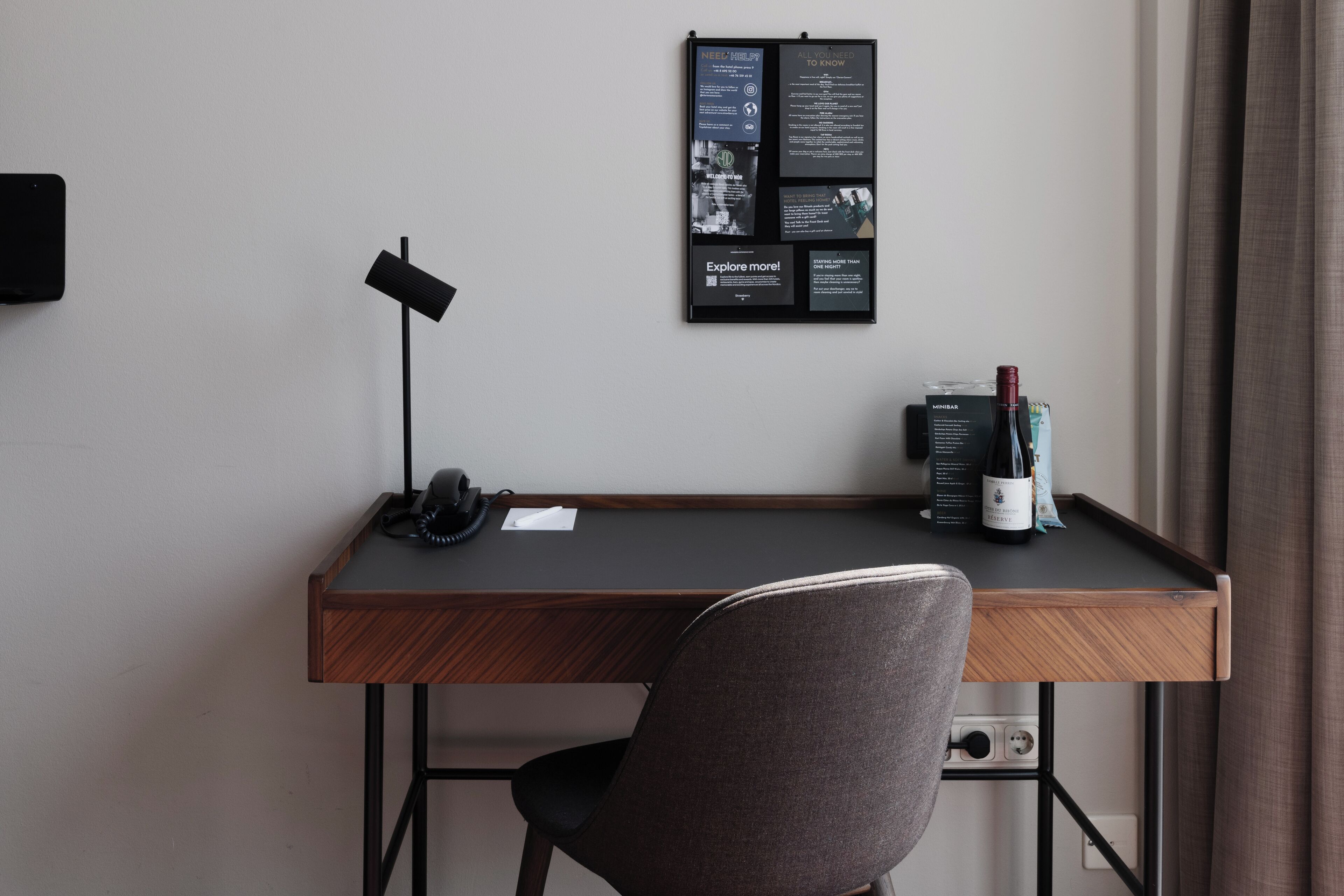 A modern hotel room with a wooden desk, a chair, a desk lamp, a wine bottle, and a bulletin board with information.