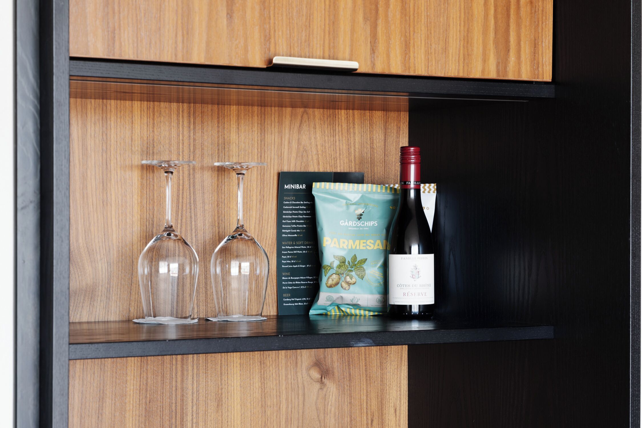 A wooden shelf with two wine glasses, a wine bottle, and a packet of Parmesano snacks.