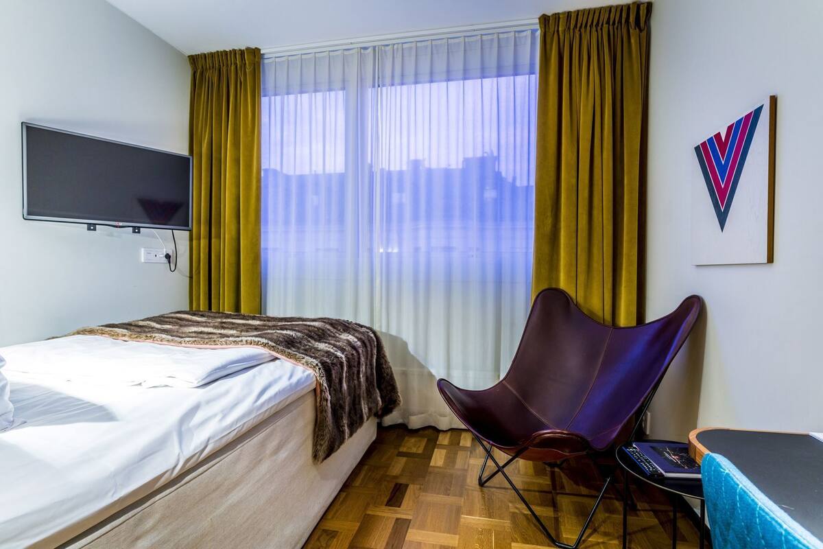 Quarto de hotel com cama, poltrona, escrivaninha, TV de tela plana e janela com cortinas.