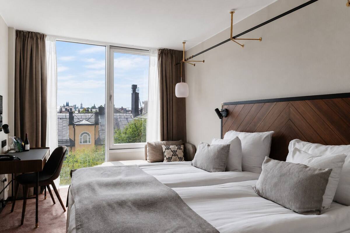 Quarto de hotel com uma janela grande, uma escrivaninha e uma cama com vários travesseiros.