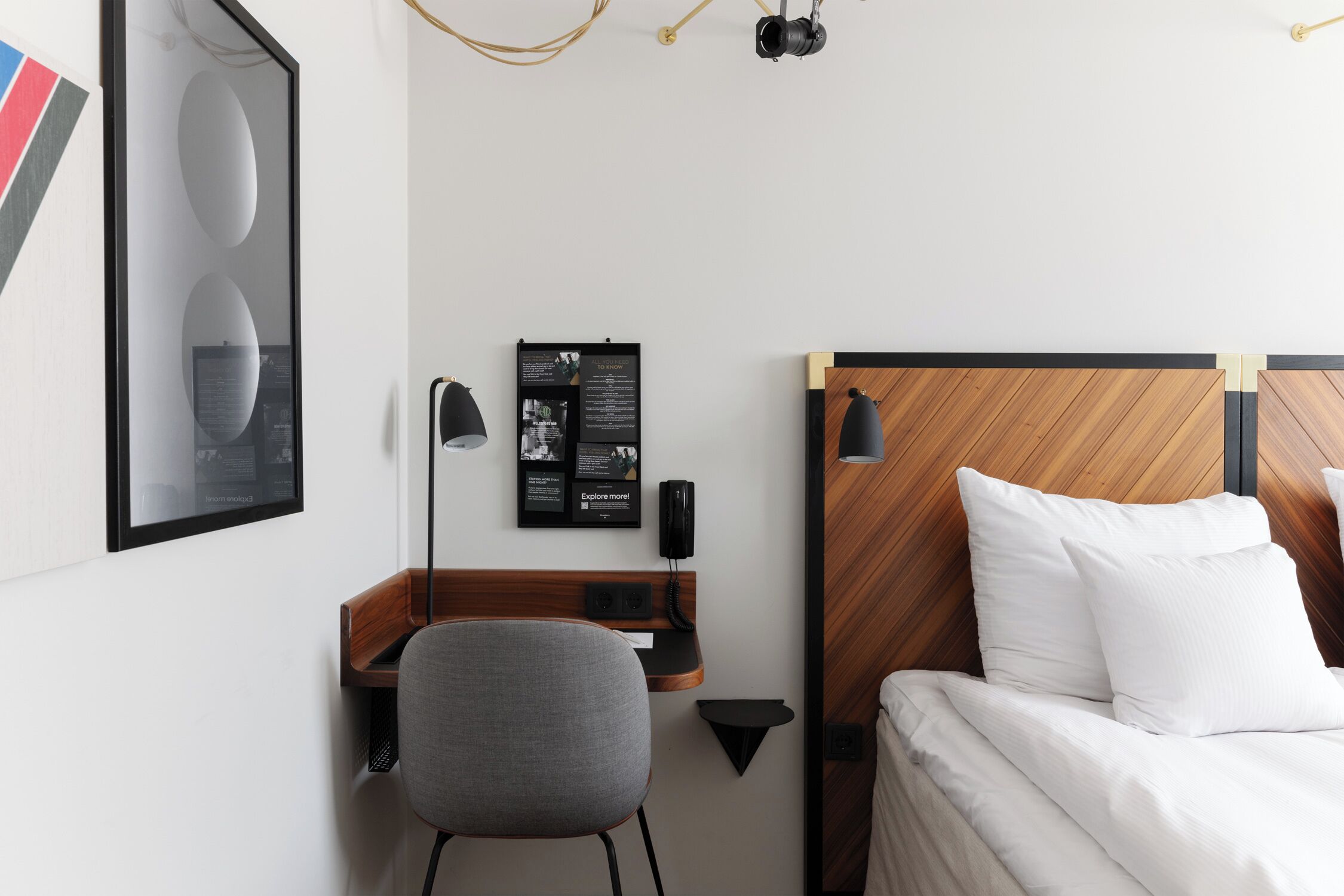 Et hotelværelse med en træsenggavl, et skrivebord med stol, et indrammet billede, en sort telefon og en vægmonteret lampe.