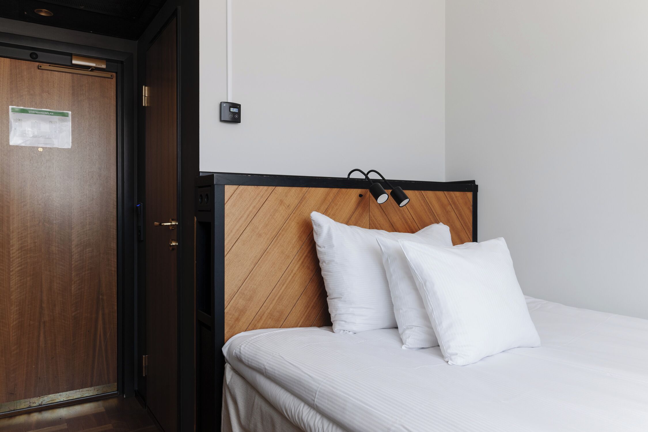 Ein Hotelzimmer mit Holz-Kopfteil, weißer Bettwäsche und einem sichtbaren Thermostat an der Wand.