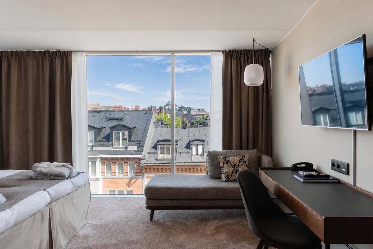 Quarto de hotel com cama, escrivaninha, cadeira, TV e uma grande janela com vista para a cidade.