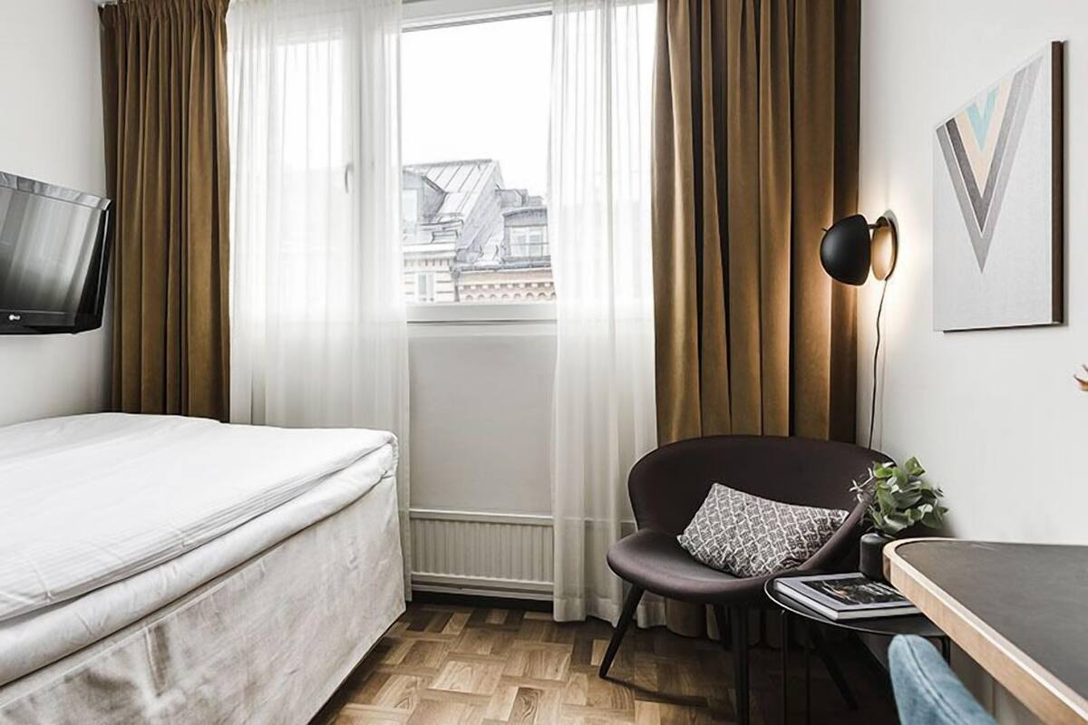 Quarto de hotel com cama, escrivaninha com cadeira, televisão fixada na parede e janela com cortinas.