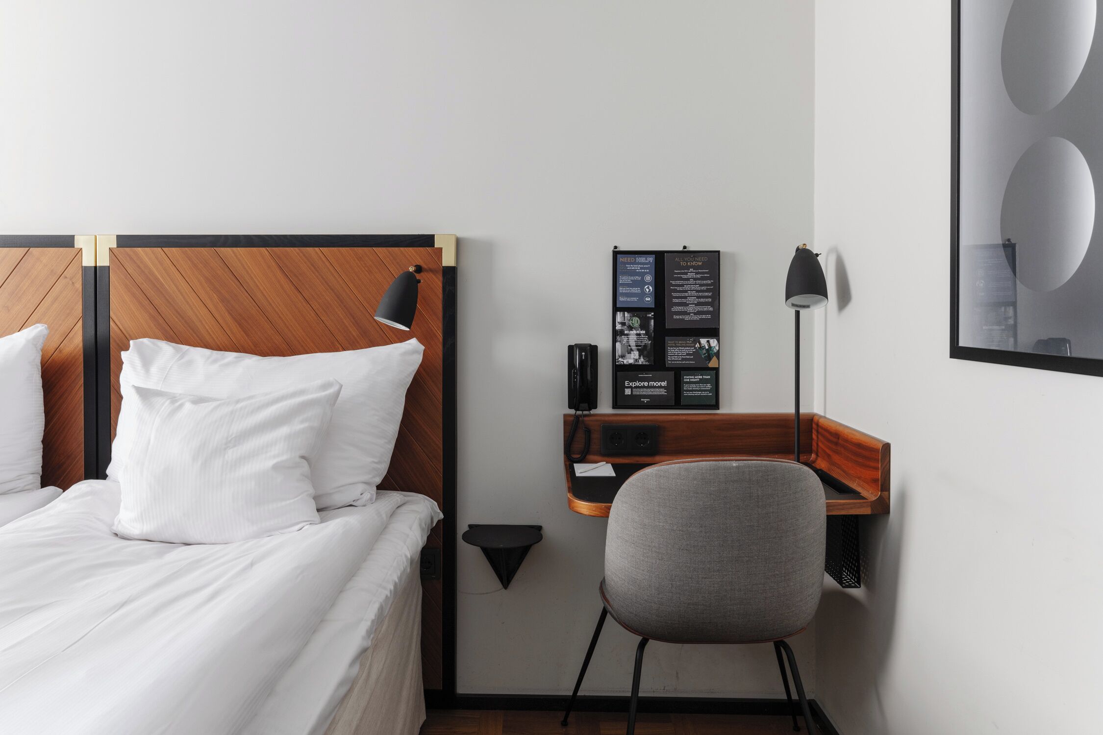 Quarto de hotel com cabeceira de madeira, uma escrivaninha com luminária, uma cadeira e um mural com informações.