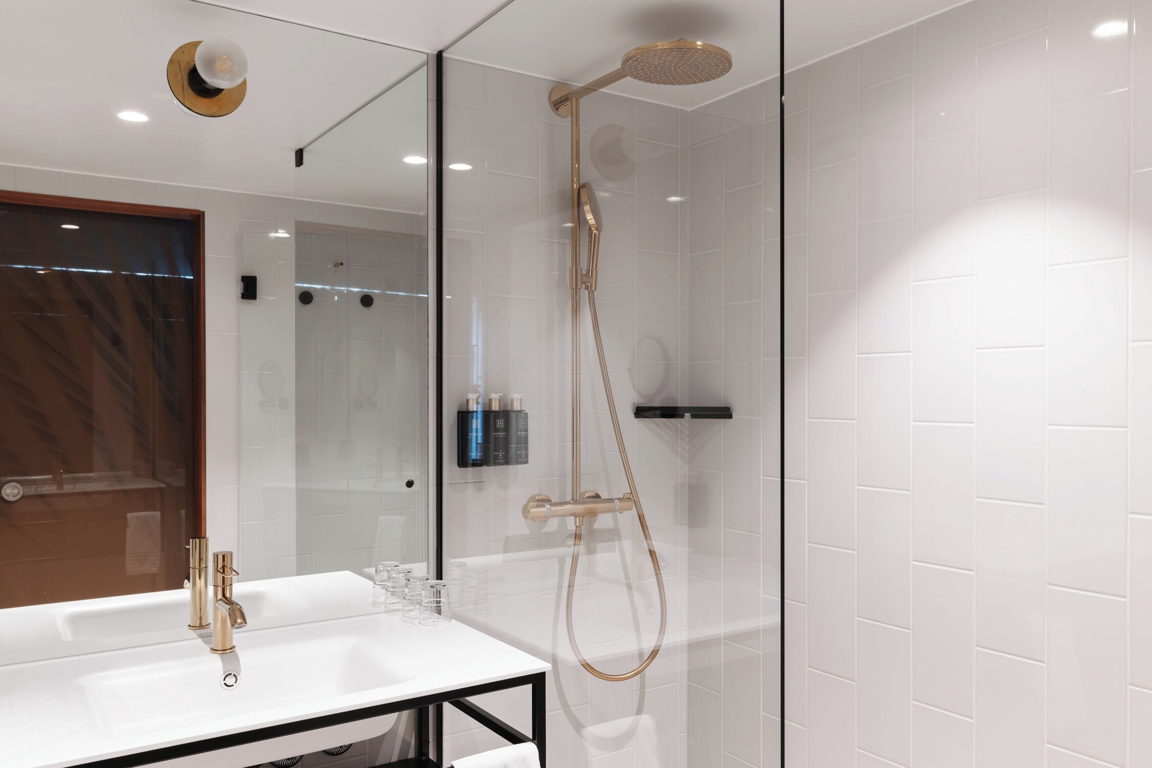 Un baño moderno con ducha acristalada, un lavabo blanco con detalles dorados y un espejo.