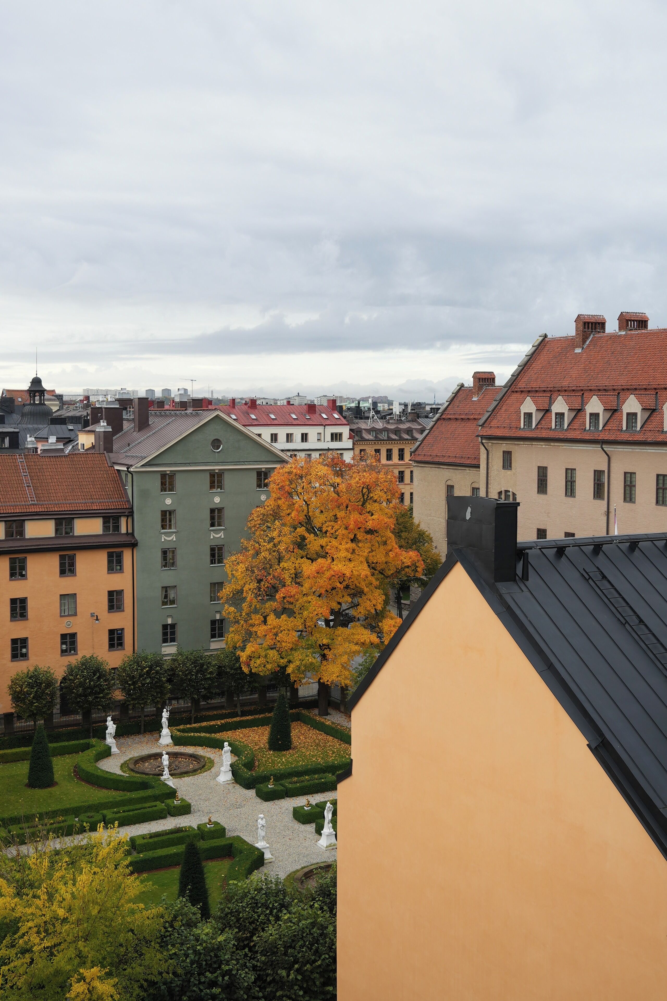 Blick auf eine Stadtlandschaft mit Gebäuden, ein Garten mit Statuen und ein Baum mit Herbstlaub.