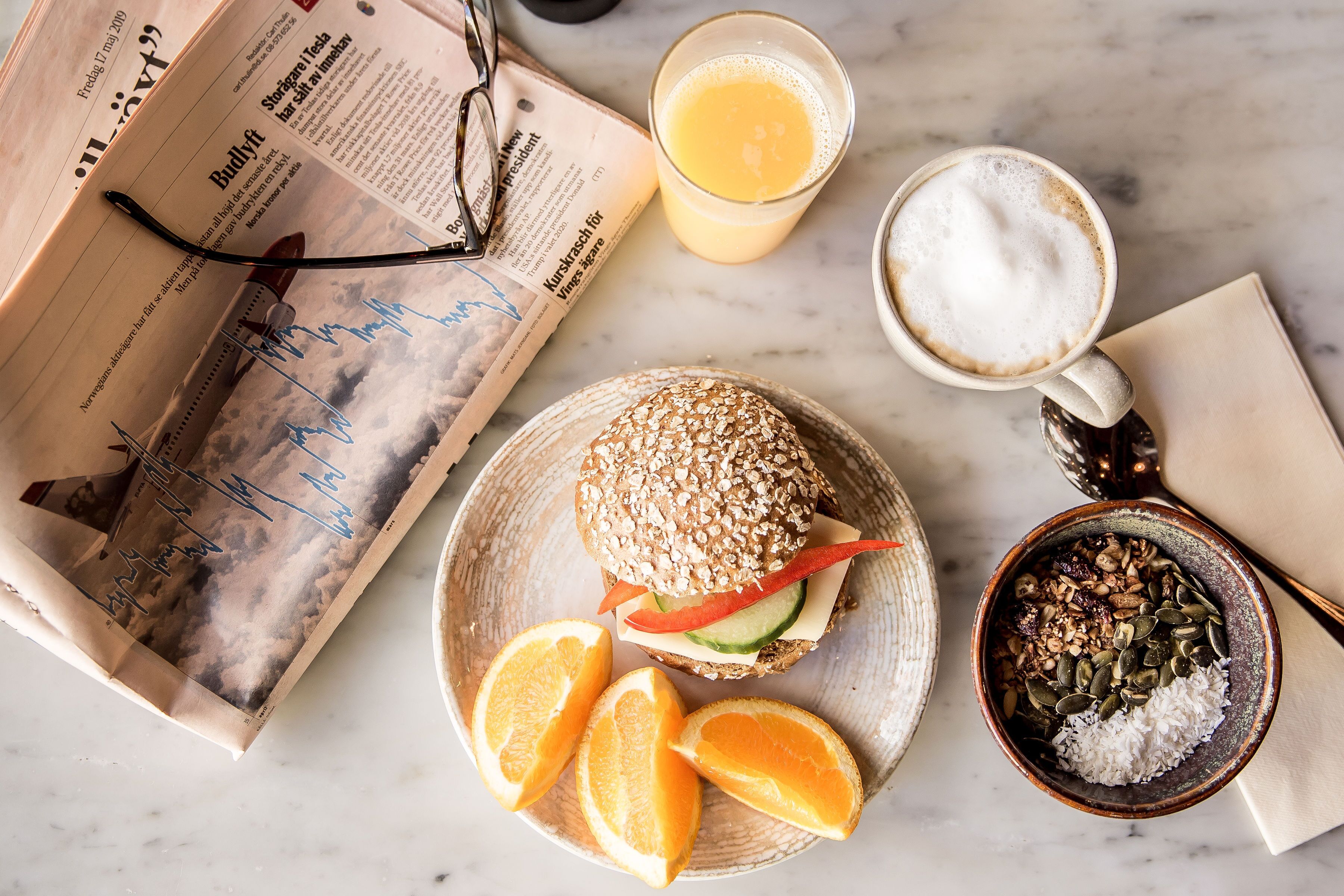 Een maaltijd bestaande uit een sandwich, sinaasappelschijfjes, een kom granola en een glas sinaasappelsap op een marmeren oppervlak.