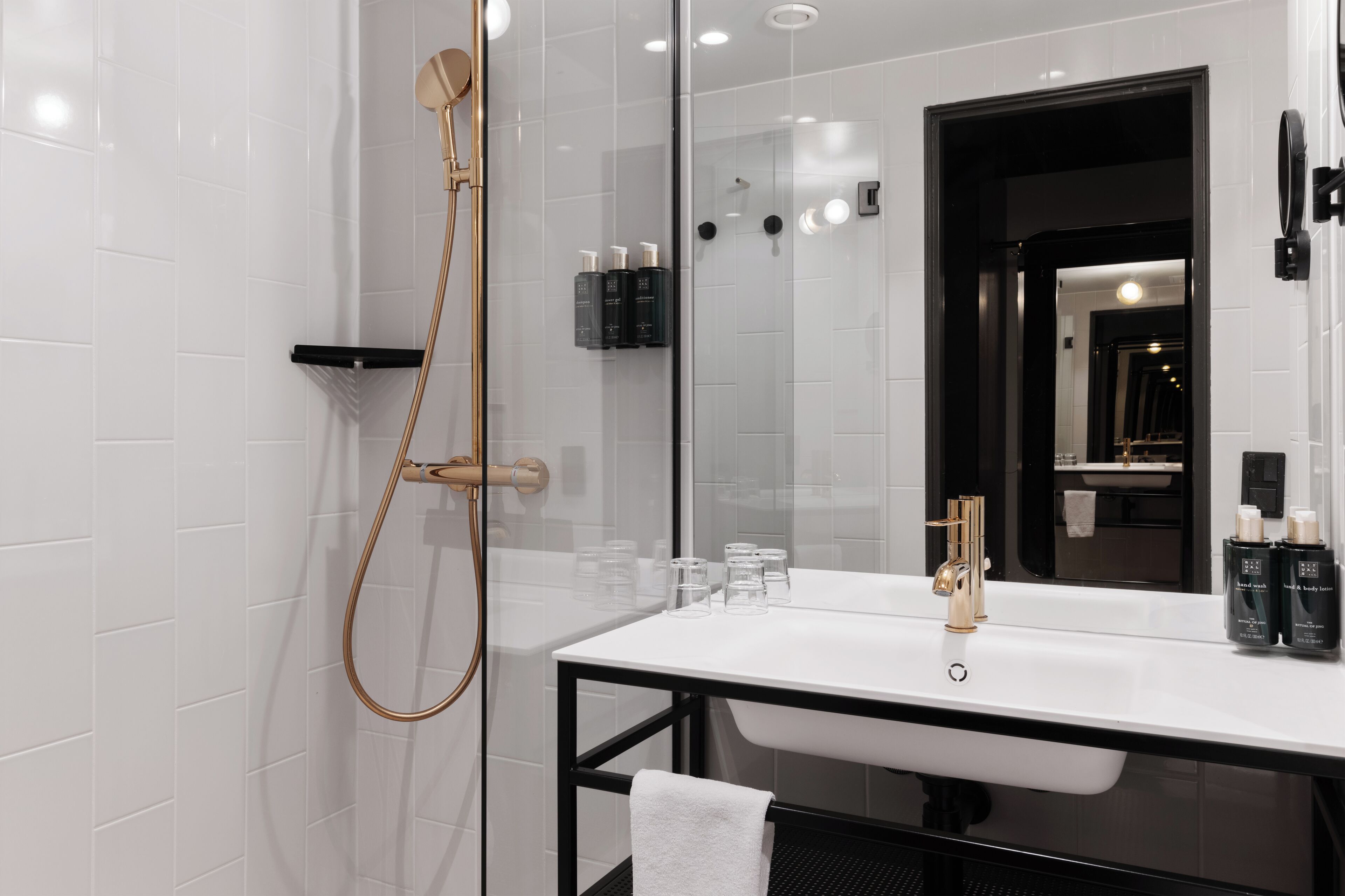 Un baño moderno con ducha envuelta en vidrio, un lavamanos blanco con detalles dorados y un espejo con marco negro.
