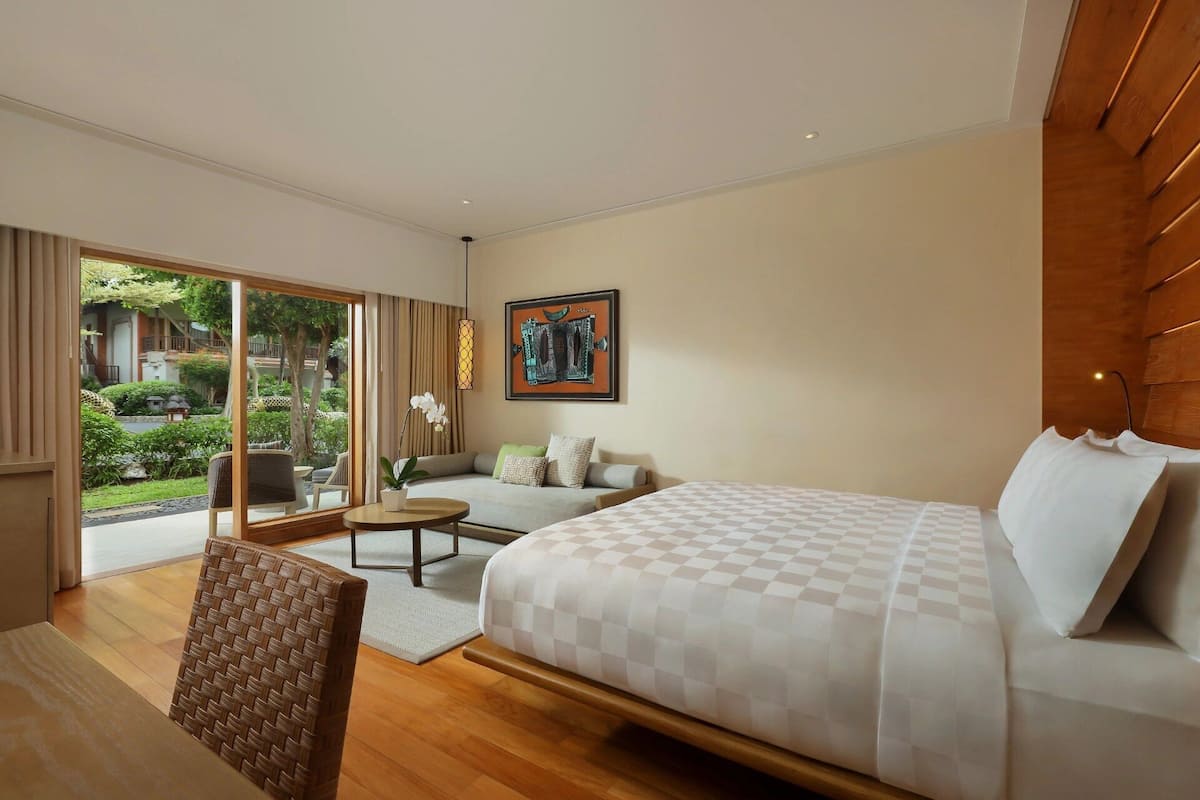 A hotel room with a large bed, a sofa, a dining table, and a view of a garden through the sliding glass door.