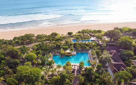 An aerial view of a resort with a central swimming pool surrounded by lush greenery and palm trees, overlooking a beach and ocean.