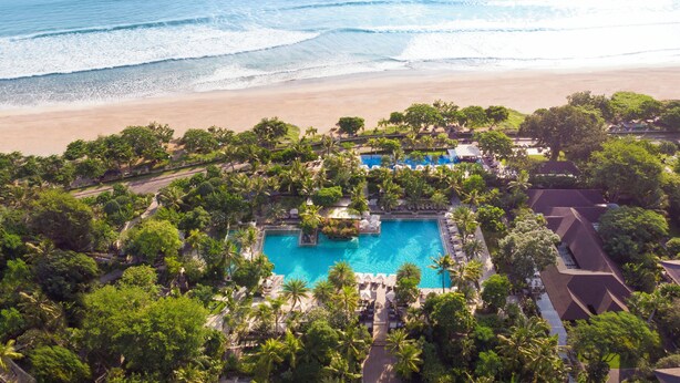 Vista aérea de un resort con piscina central rodeada de exuberante vegetación y palmeras, con vistas a una playa y al océano.