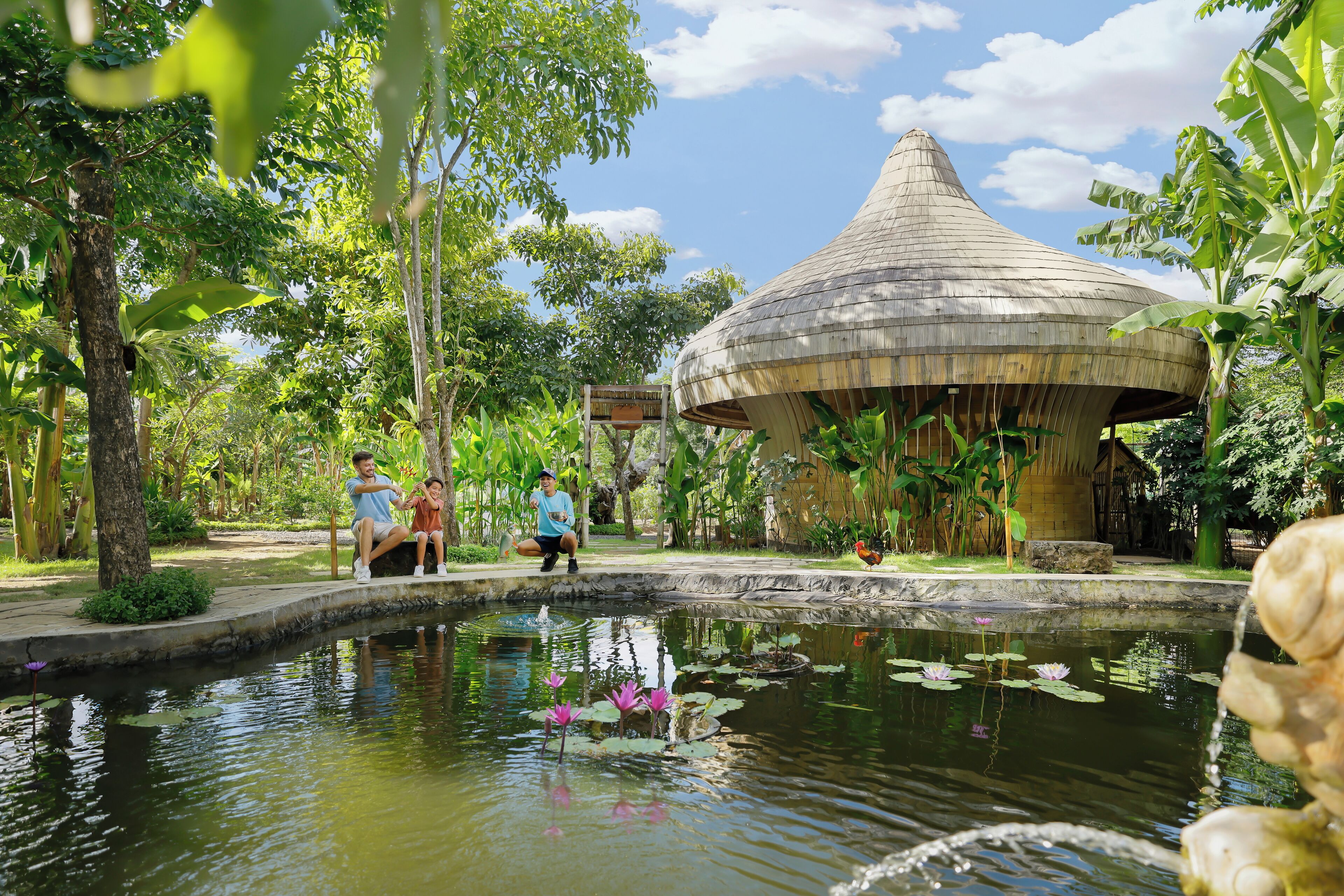A traditional thatched-roof pavilion surrounded by a lush garden with a pond featuring lotus flowers.