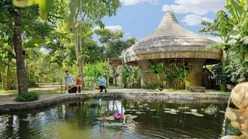 A traditional thatched-roof pavilion surrounded by a lush garden with a pond featuring lotus flowers.