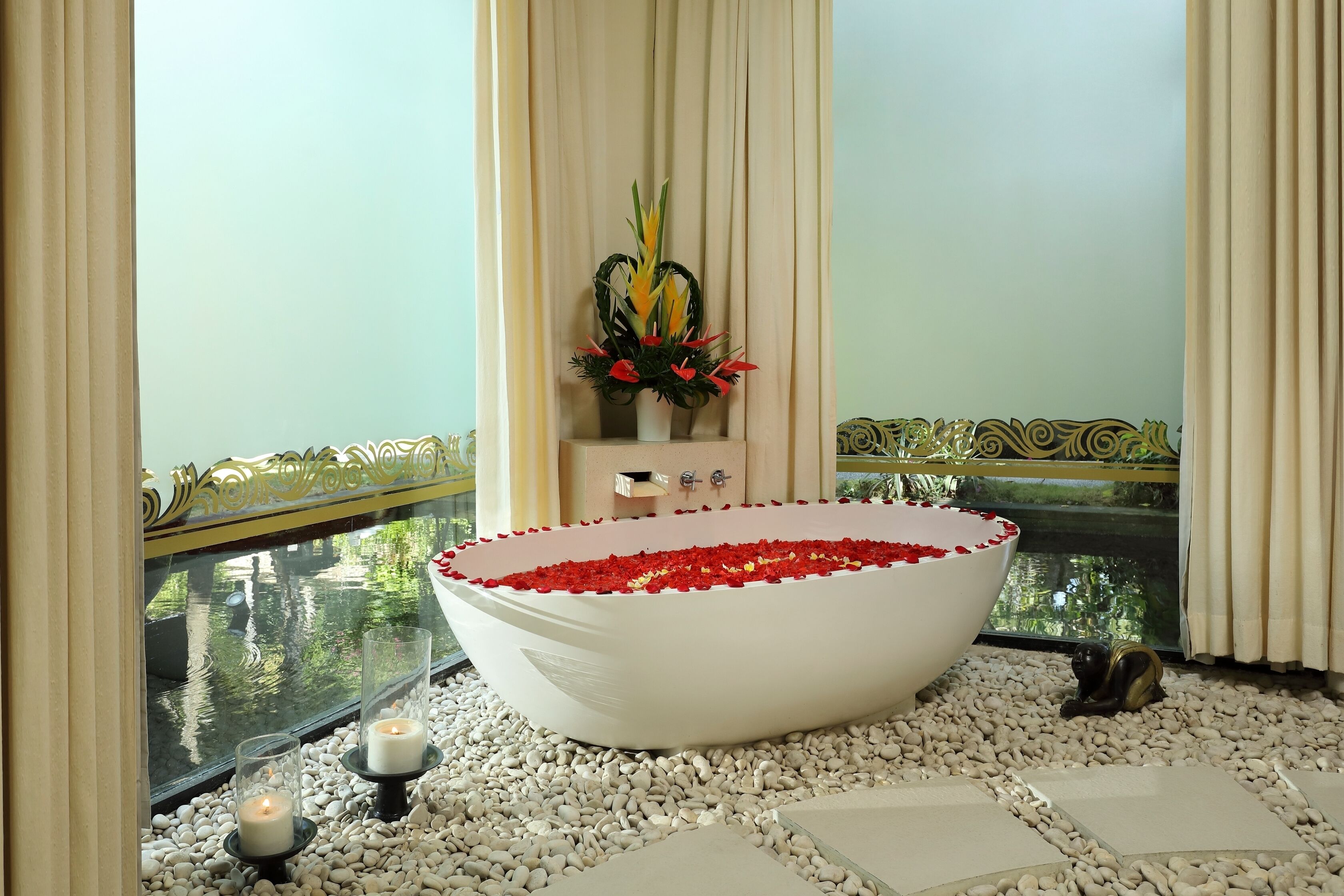 Una bañera independiente grande llena de pétalos de rosa, rodeada de guijarros blancos, con velas y una planta decorativa en un balcón con vistas a un cuerpo de agua.