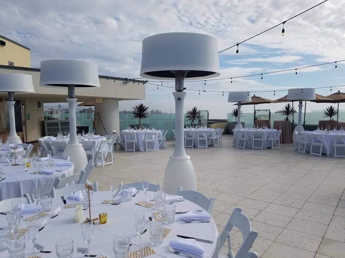 Un espace de restauration en plein air avec des tables rondes dressées pour un repas, entourées de chaises blanches et de grandes lampes blanches.
