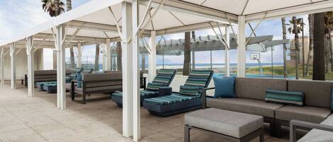 A covered outdoor seating area with lounge chairs and sofas, a basketball hoop, and a view of the beach.
