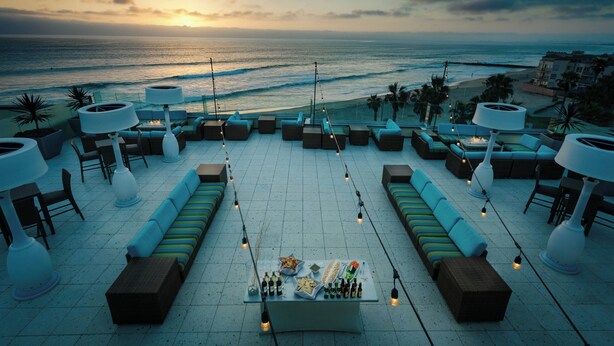 A rooftop terrace with seating, a table with bottles, and a view of the ocean at sunset. - Pier South Resort, Autograph Collection by Marriott (Imperial Beach)