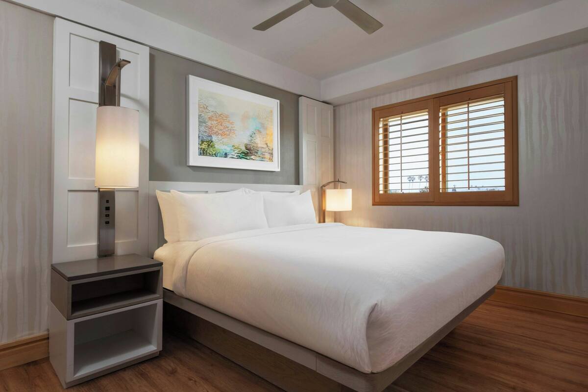 Camera d'albergo ordinata con un letto, comodini, una ventola a soffitto, una finestra con tende e un quadro appeso al muro.