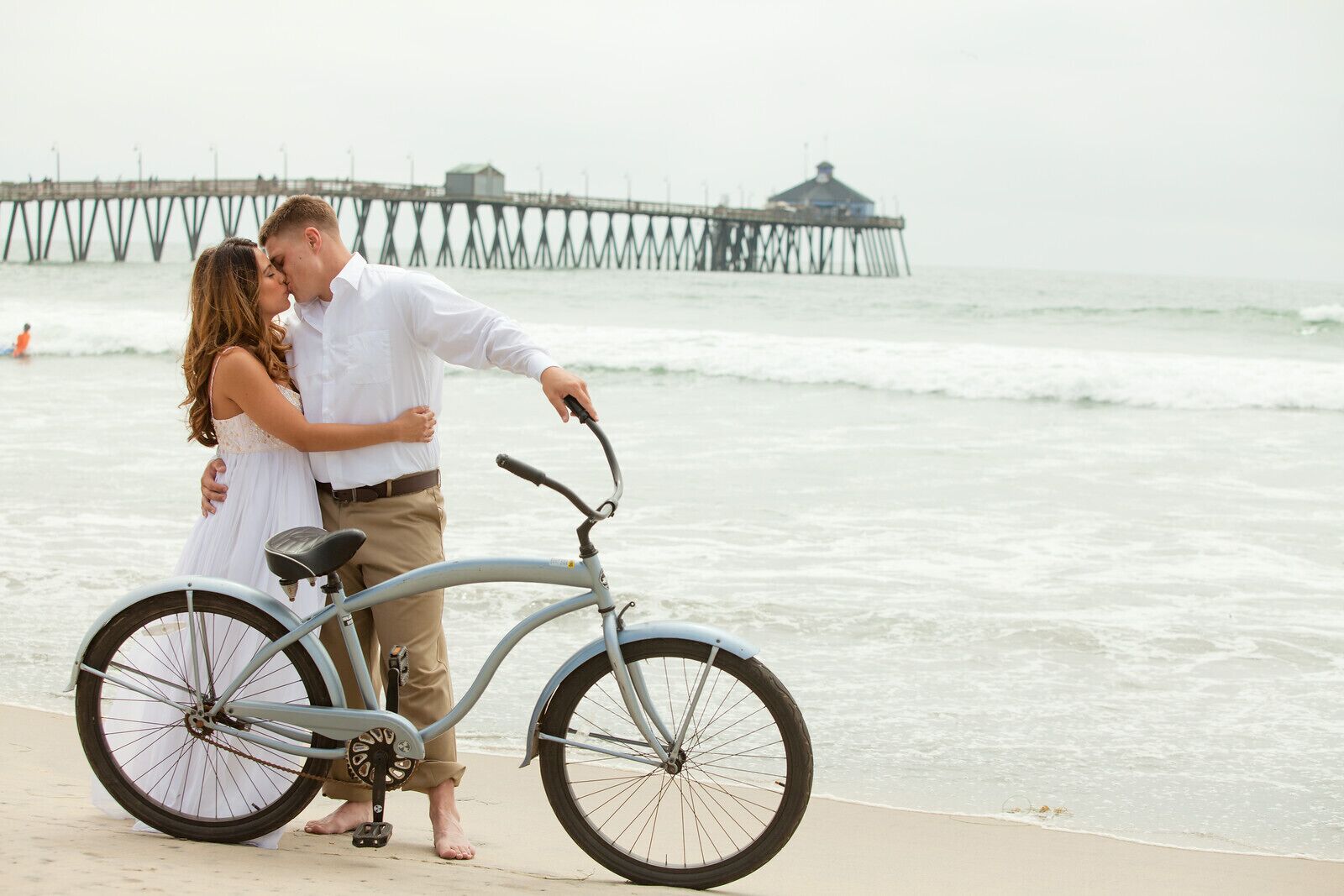 A couple embracing near a bicycle on a beach with a pier in the background.