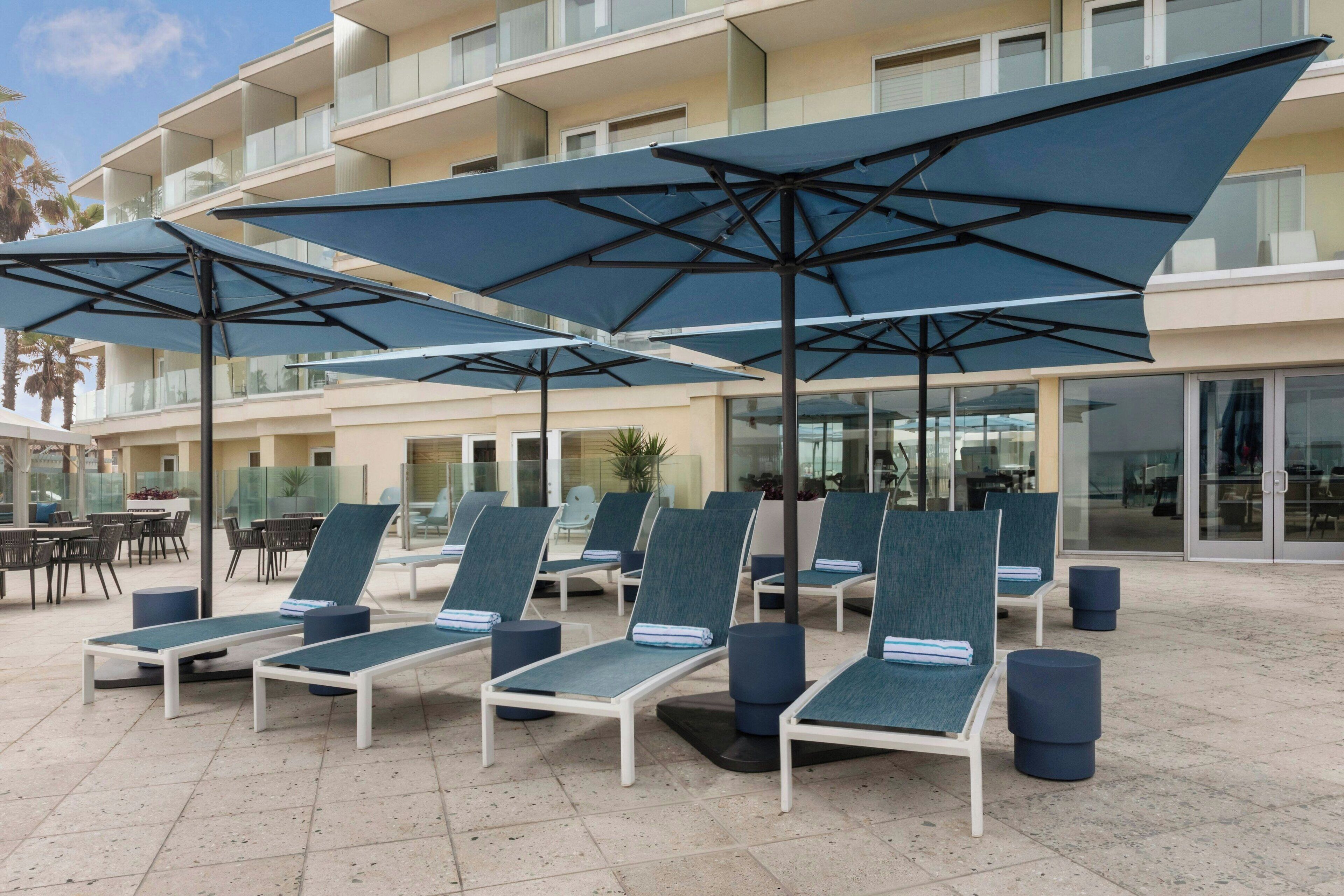 Un área junto a la piscina con sillones de baño y sombrillas, un edificio con balcones y mobiliario de exterior.
