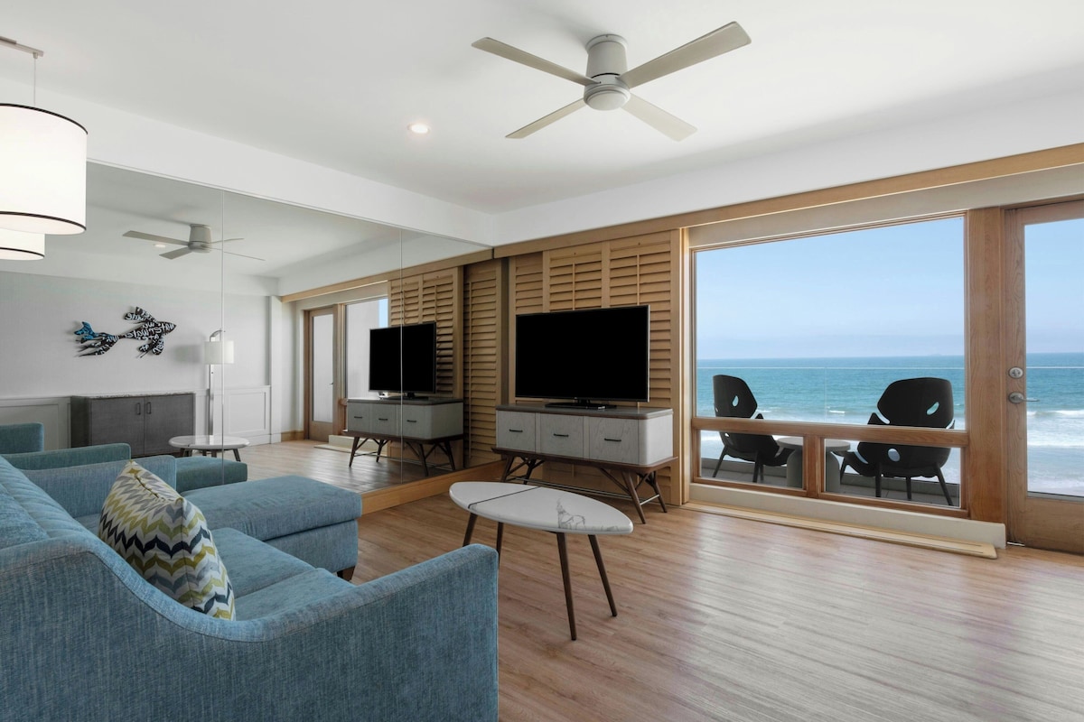 A modern living room with a large TV, comfortable seating, and a view of the ocean.