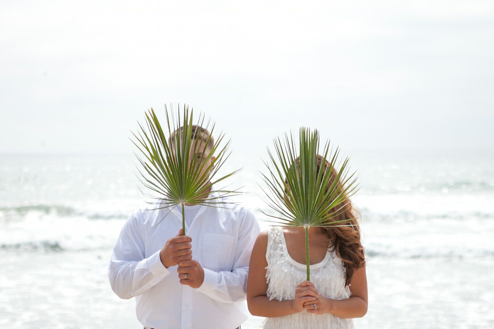 Un couple tenant des palmes devant leur visage, sur la plage.