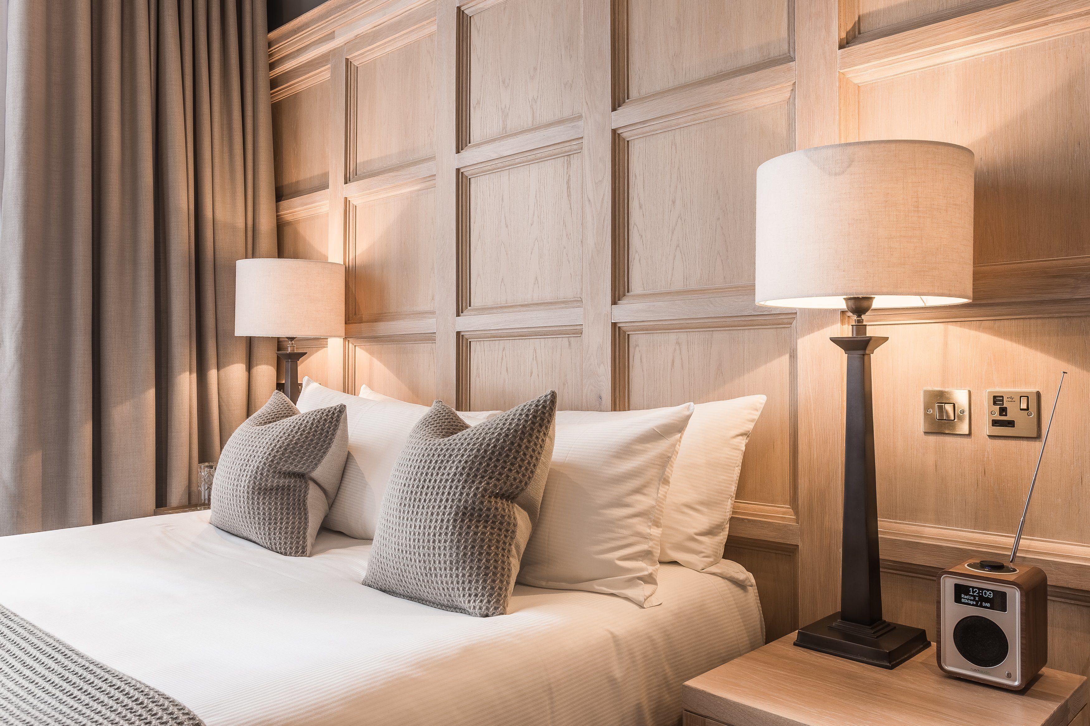 Habitación de hotel con una cama grande, dos lámparas de noche, una radio y un interruptor de luz en la pared.