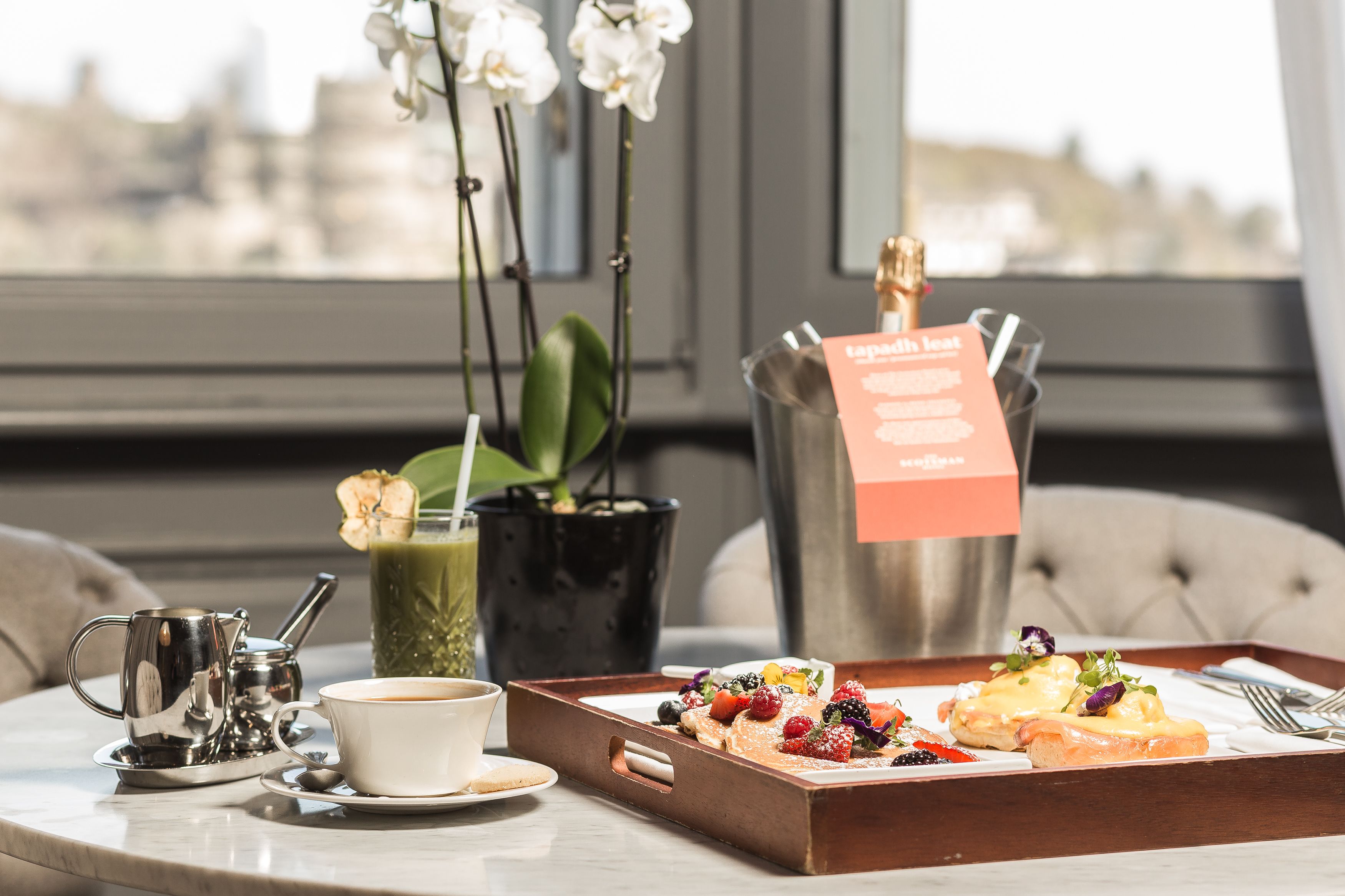 Un plateau de petit-déjeuner avec des crêpes, des fruits rouges et une bouteille de champagne, une tasse à café, une théière et une plante en pot, le tout posé sur une table dans une pièce offrant une vue sur la ville.