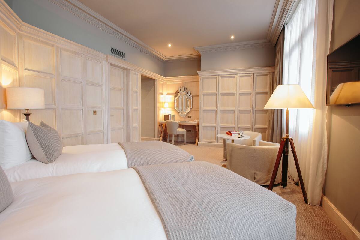 A hotel room with two beds, a desk, a chair, a lamp, and a mirror.