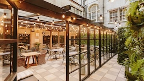 A covered outdoor seating area with tables and chairs, surrounded by glass walls and a brick building in the background.