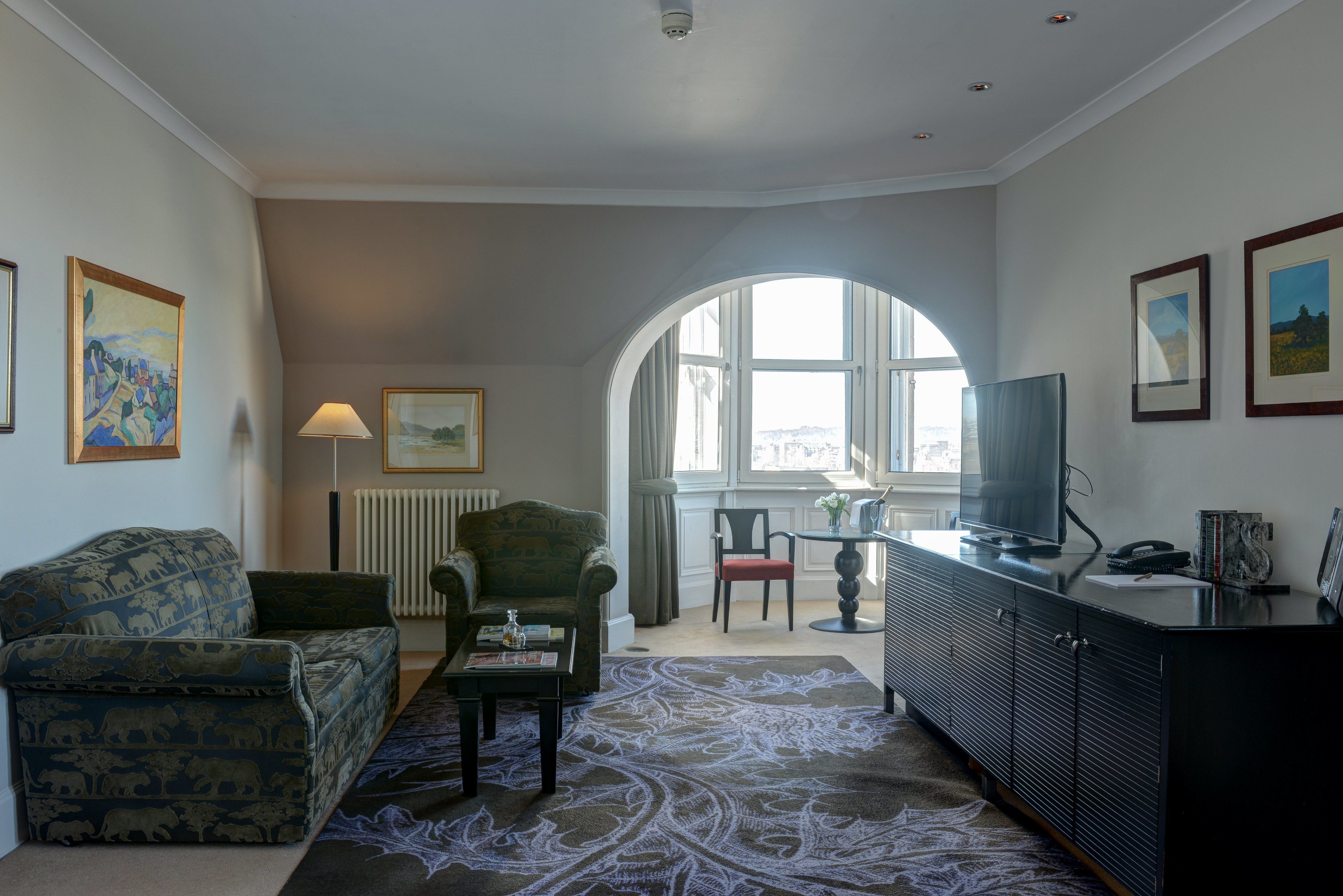 Een woonkamer met een bank, fauteuil, salontafel en een televisie op een kast. Er is een groot raam met uitzicht op de stad.