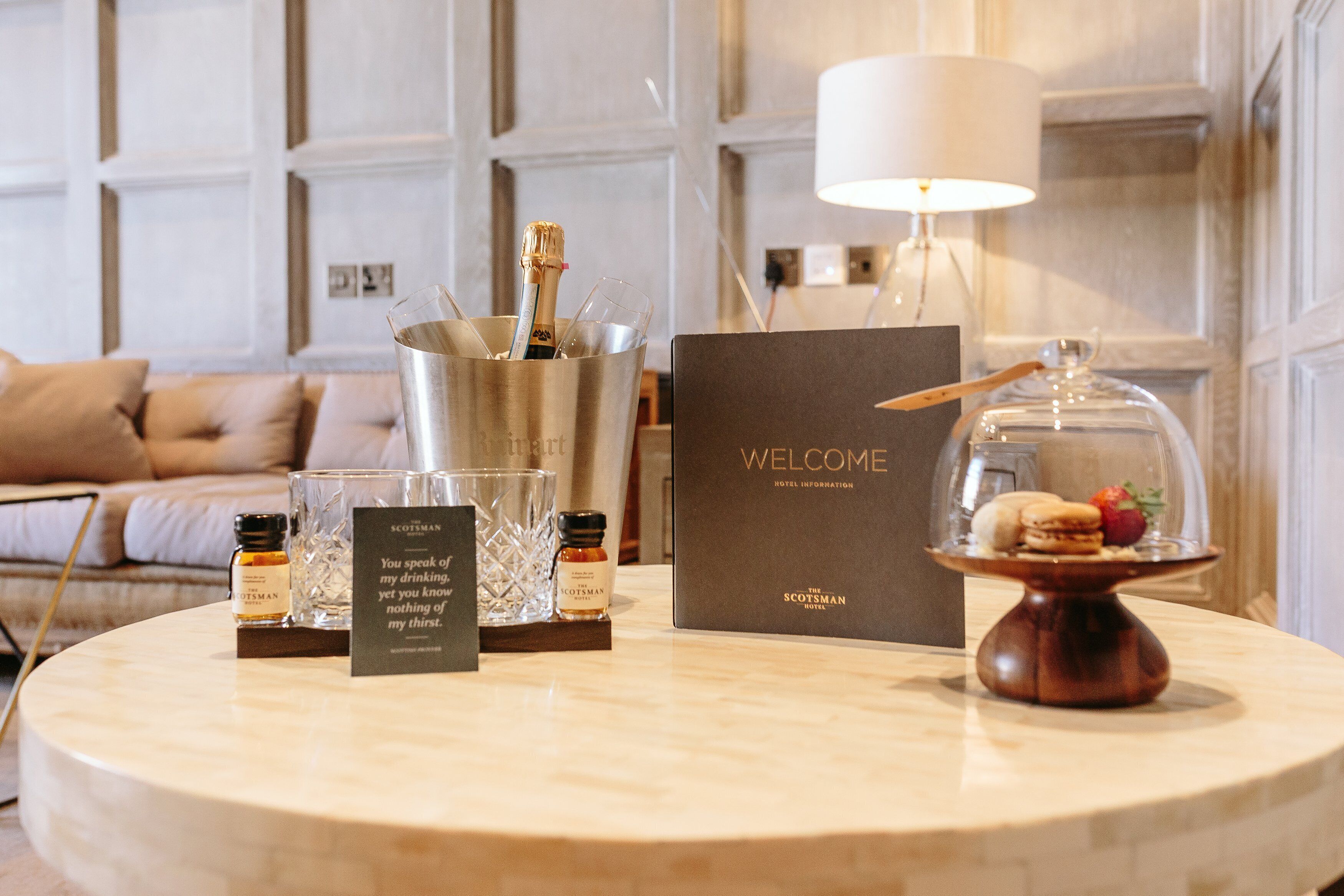 A hotel reception area with a welcome box, a bottle of Scotchman whisky, a glass of whisky, and a dessert display.