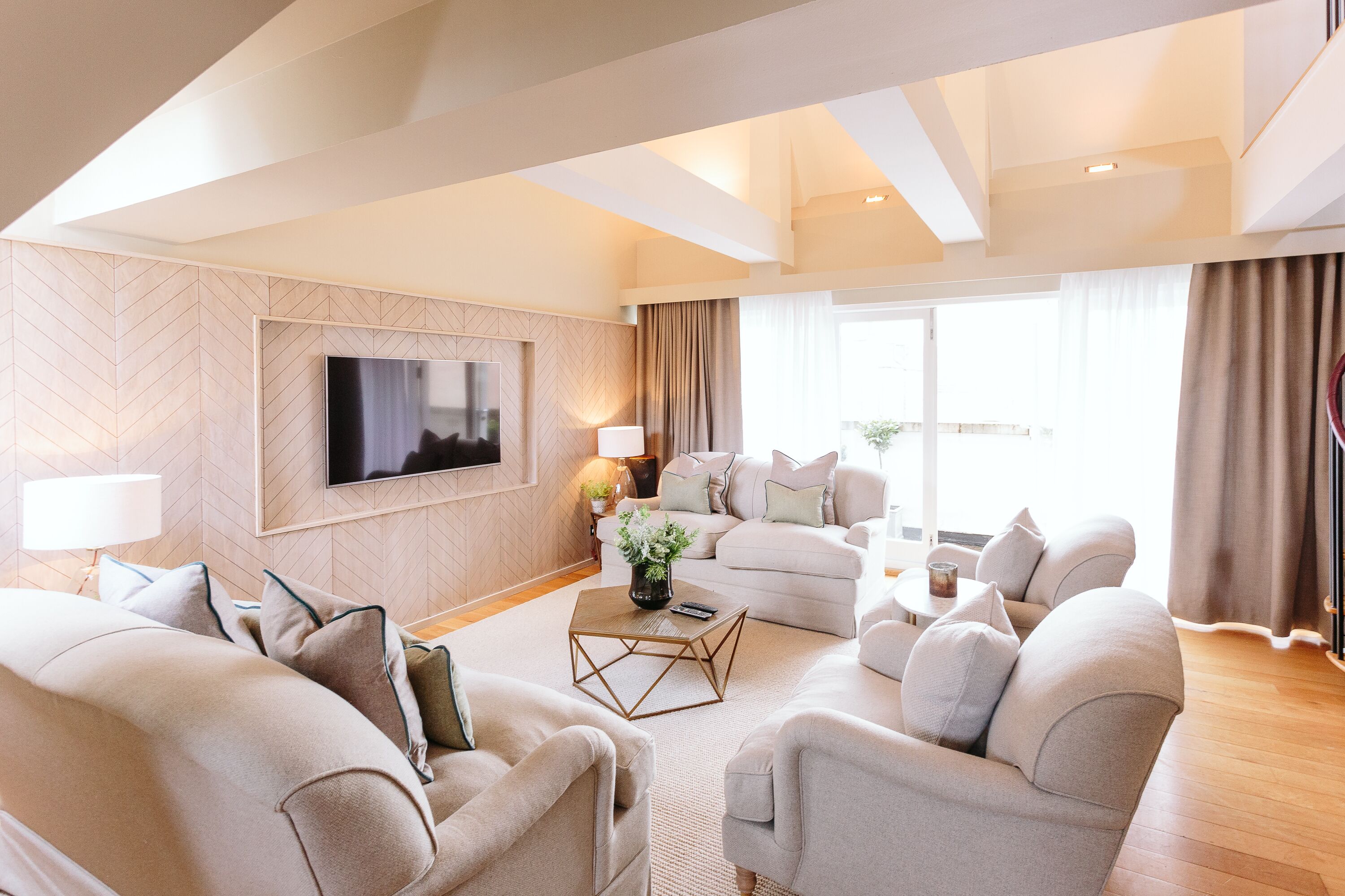 Un salon moderne avec un grand téléviseur à écran plat fixé au mur, un ensemble de canapés de couleur beige, une table basse et une grande fenêtre avec des rideaux.