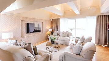 A modern living room with a large flat-screen TV mounted on the wall, a beige sofa set, a coffee table, and a large window with curtains.