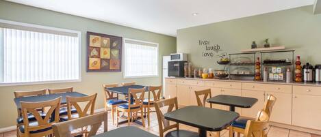 A dining area with wooden tables and chairs, a kitchen area with a microwave and a coffee station, and a wall with a positive message.
