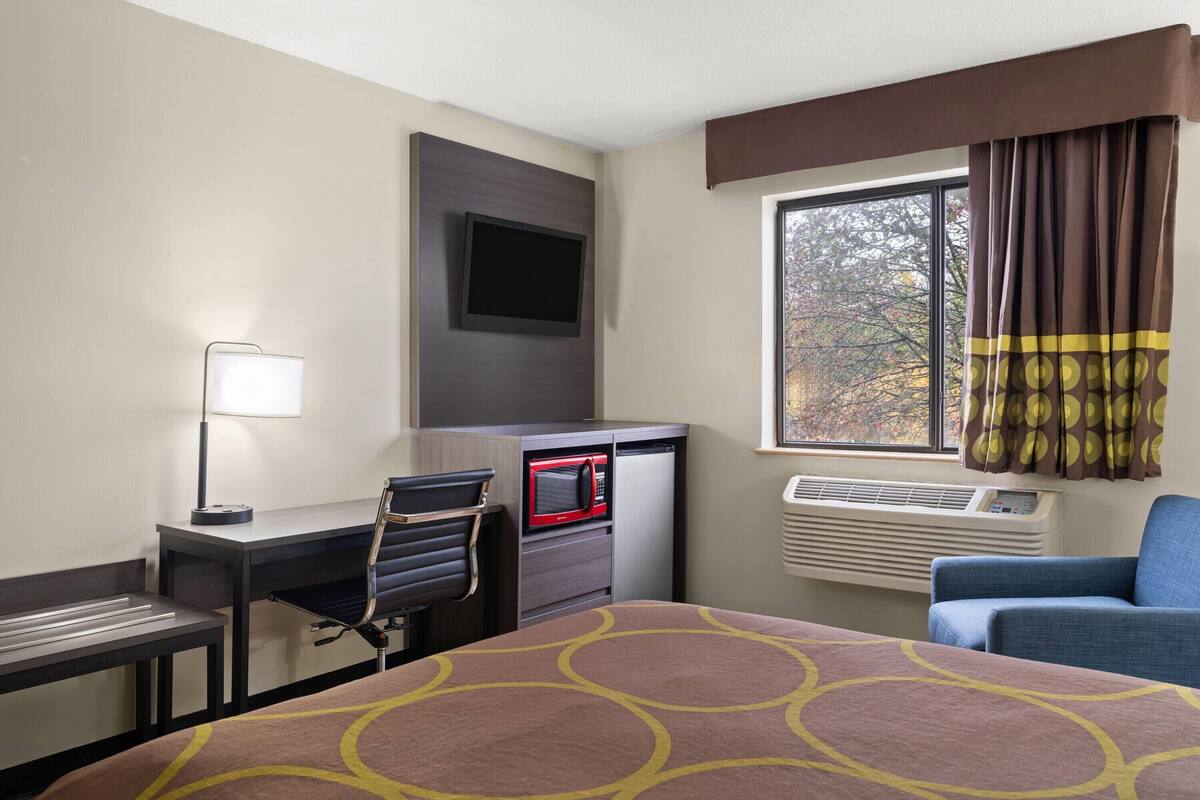 A hotel room with a bed, desk, chair, microwave, air conditioning unit, and a window with curtains.