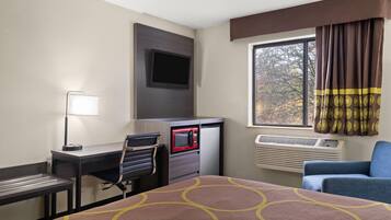 A hotel room with a bed, desk, chair, microwave, air conditioning unit, and a window with curtains.